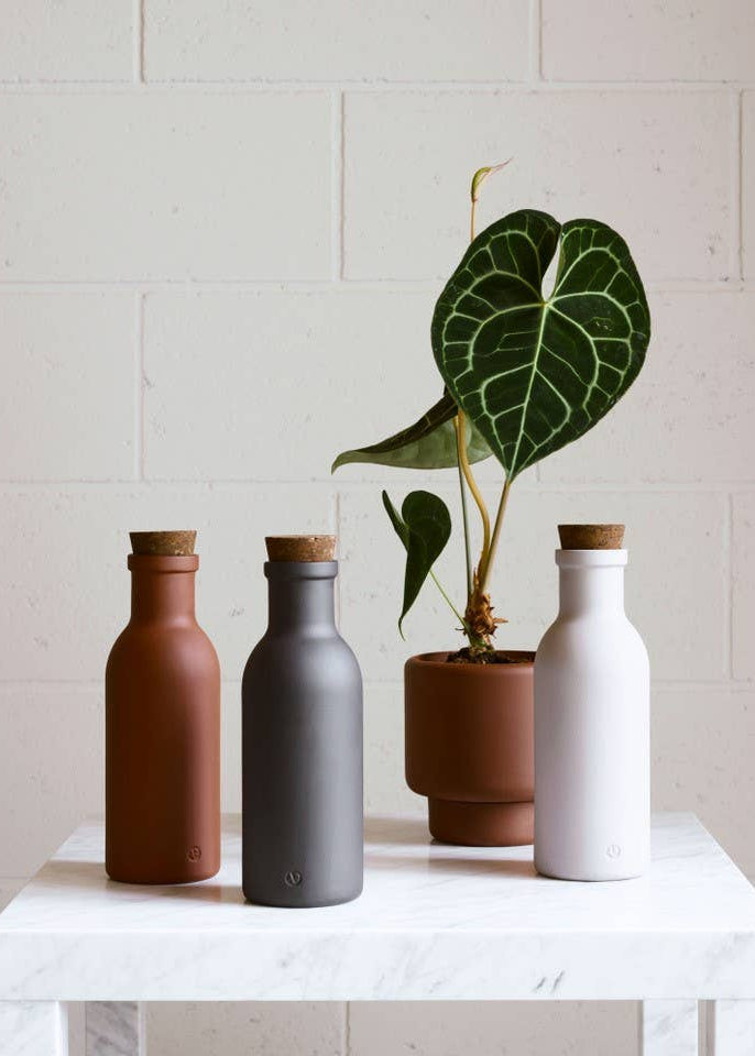 Three matte ceramic bottle-shaped planters in terracotta, charcoal gray, and off-white, arranged on a white marble side table with a potted green plant in the center, set against a light beige brick wall.
