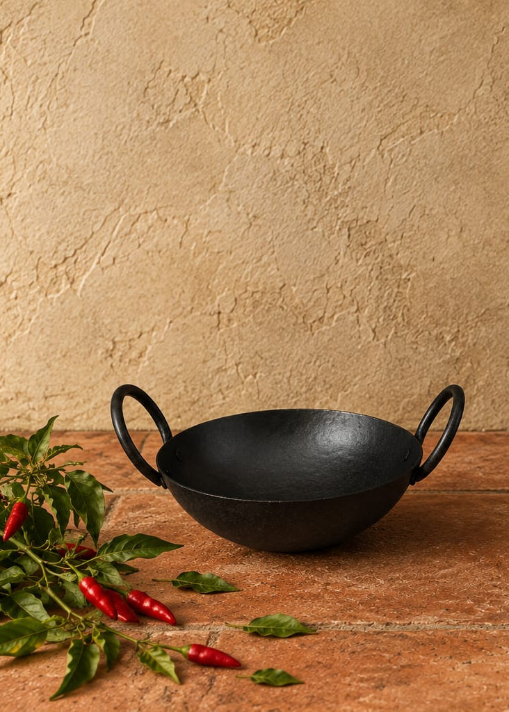 Two black cast iron kadhai pots with loop handles, displayed on a terracotta tiled surface against a textured beige stucco wall, with red chilies and green leaves as props.