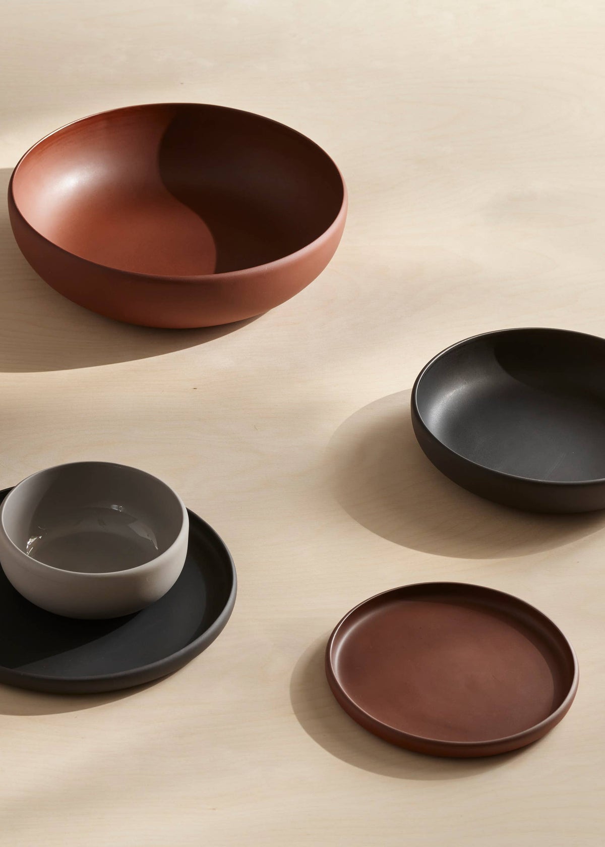A collection of matte ceramic tableware in earthy tones, including bowls, plates, and cups, arranged on a light beige wooden surface with soft natural shadows.