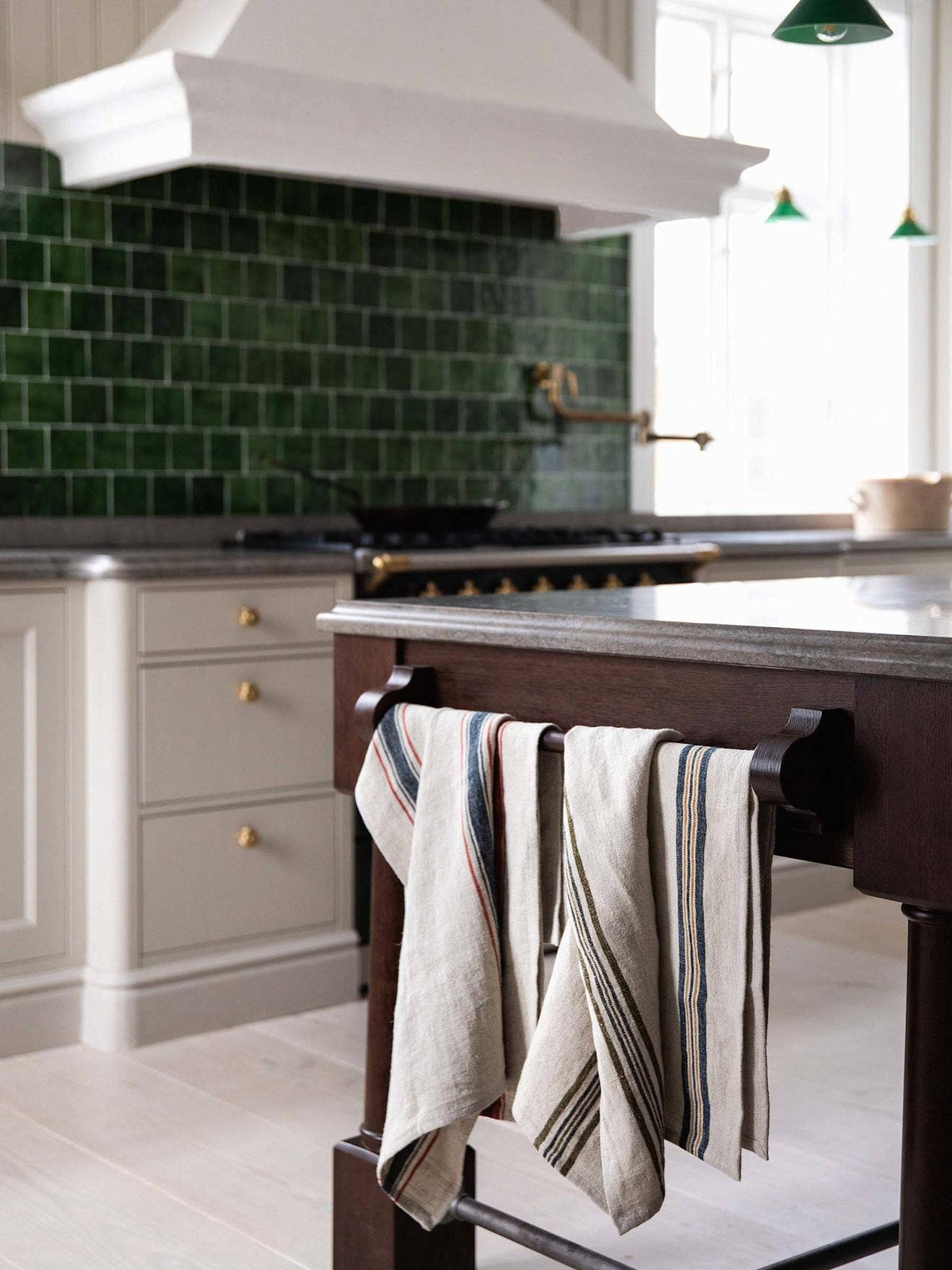 A kitchen scene with a dark wood island in the foreground holding two striped kitchen towels, set against a green subway tile backsplash and white cabinetry.