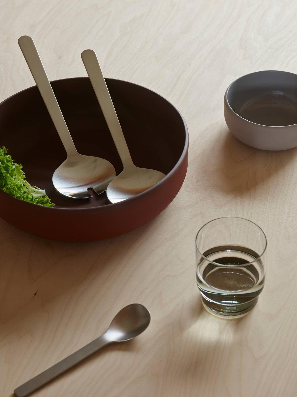 A styled tabletop scene with a dark brown bowl containing two beige-handled spoons, a small gray bowl, a clear glass of water, and a metal spoon on a light wood surface.