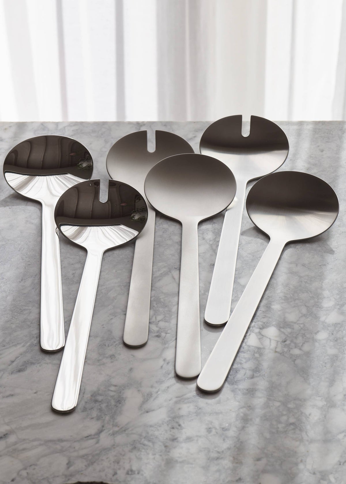 Five stainless steel serving spoons with integrated spoon bowls, arranged on a gray marble surface in front of white curtains.