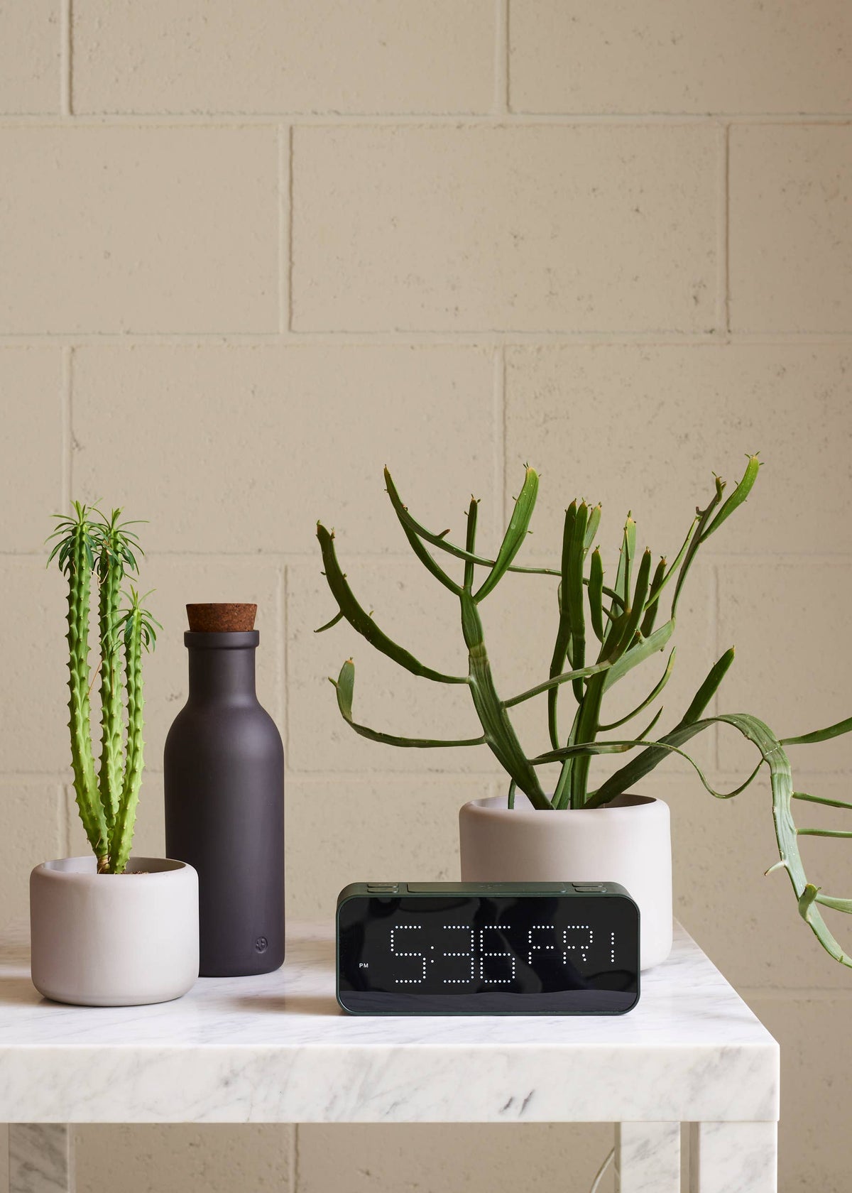 Decorative setup with potted plants and a digital clock on a marble surface against a beige brick wall.