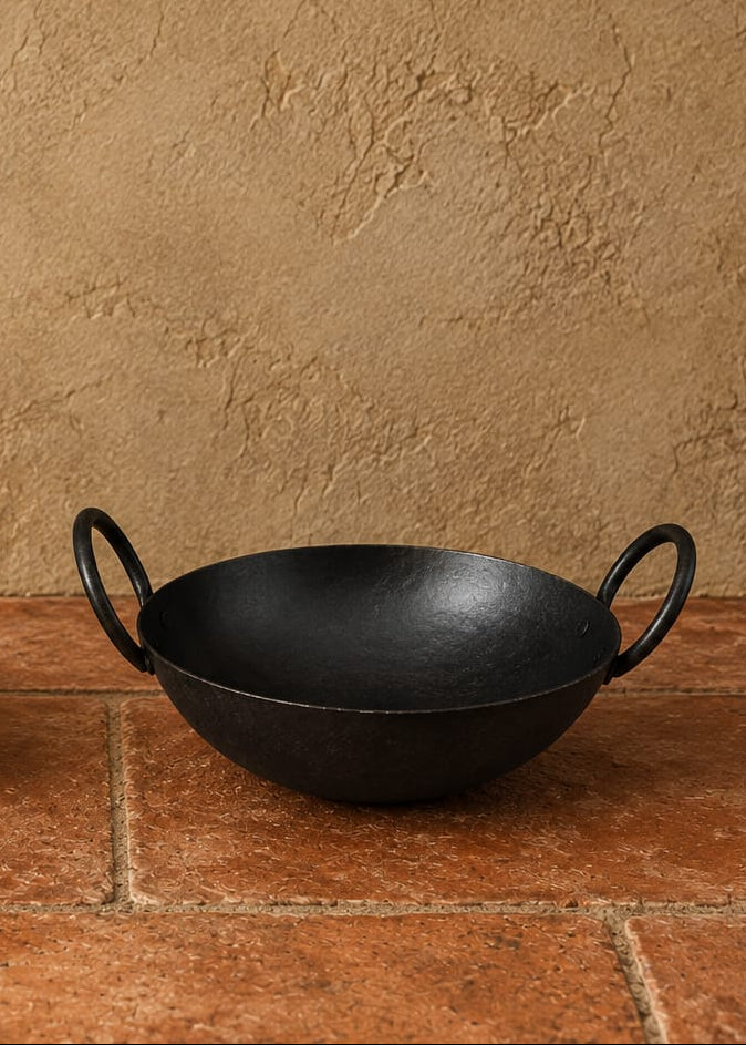 Two black cast iron kadhai pots with loop handles, displayed on a terracotta tiled surface against a textured beige stucco wall, with red chilies and green leaves as props.