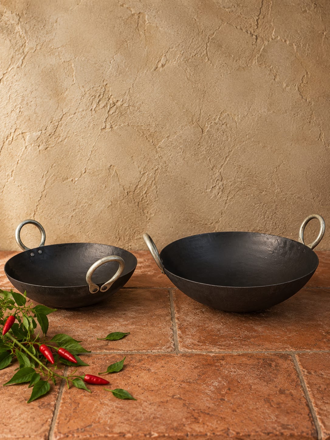 Two black cast iron kadhai pans with silver loop handles, displayed on a terracotta tiled surface against a textured beige stucco wall, with red chili peppers and green leaves as props in the foreground.