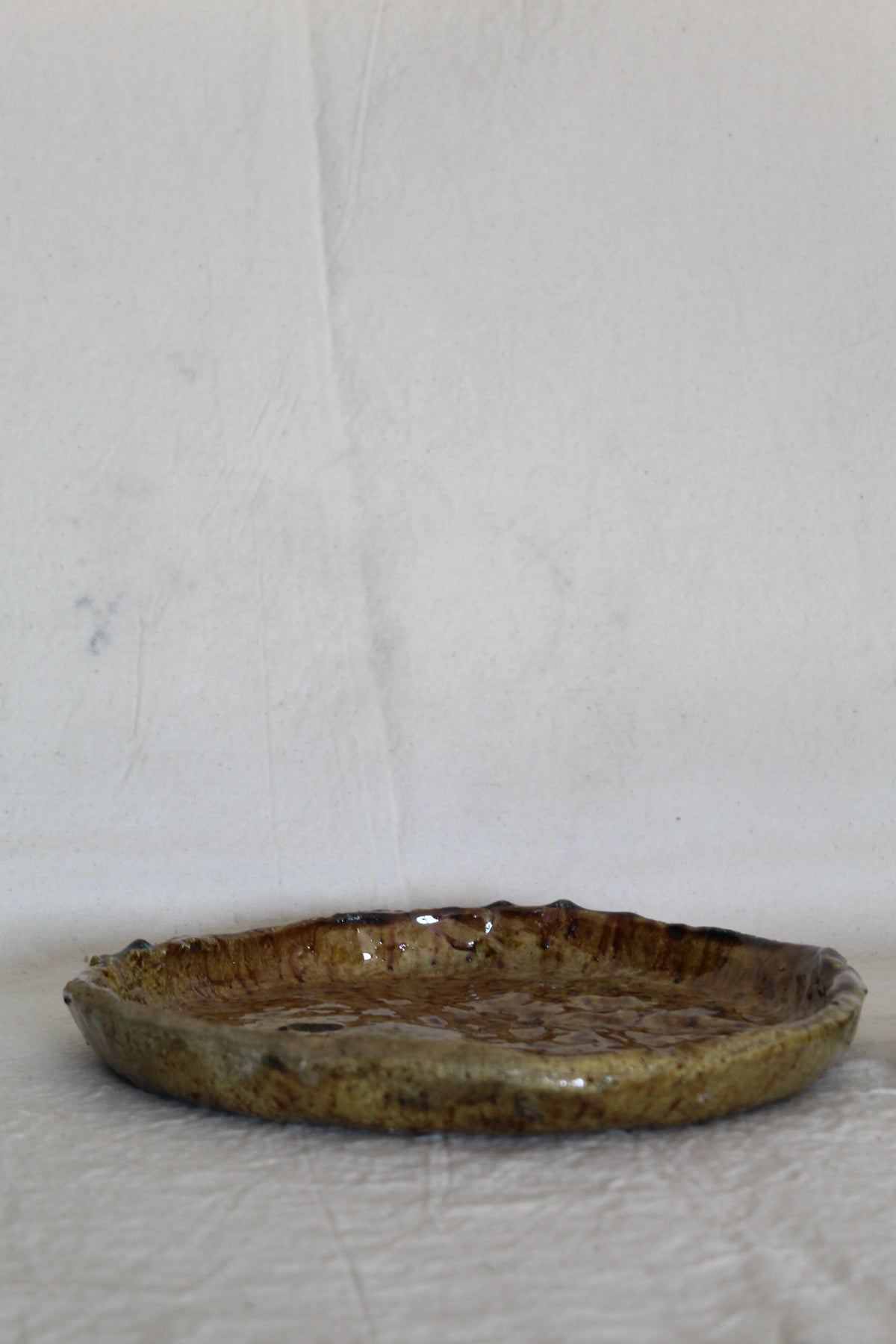 A handmade ceramic shallow dish with a rustic, hand-painted brown and gold glaze, resting on a textured white fabric surface against a white background.