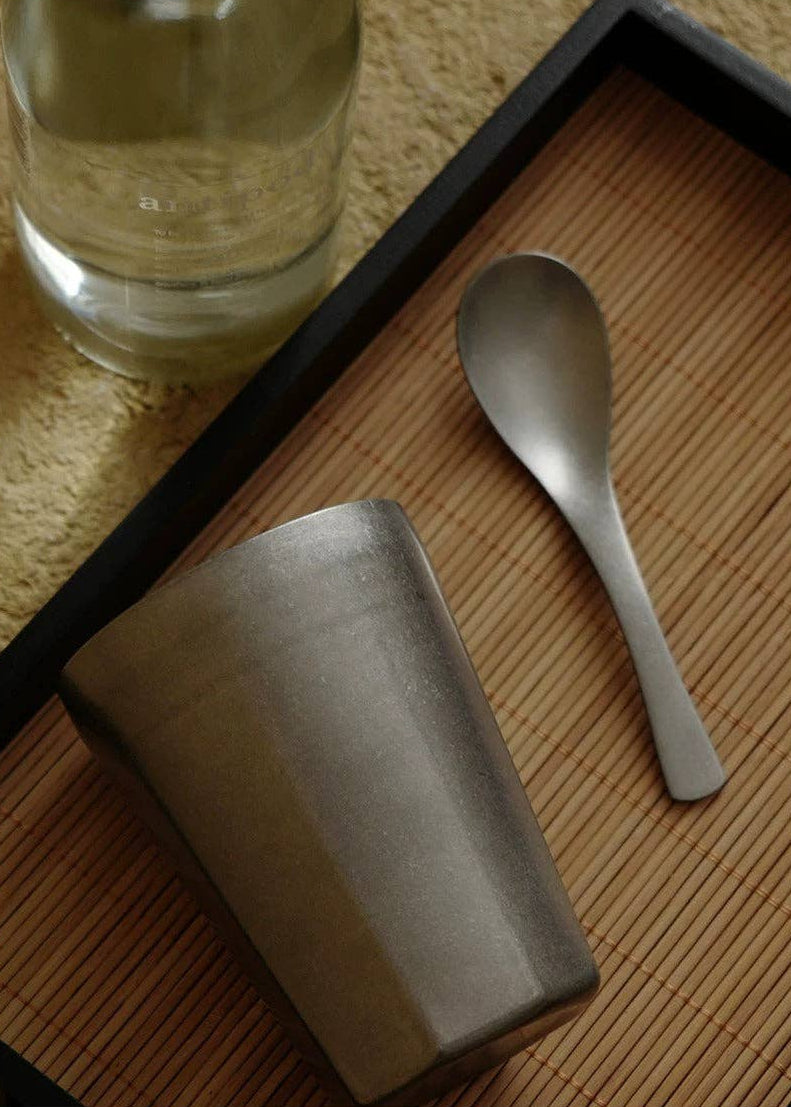 A matte gray ceramic cup and matching metal spoon resting on a brown bamboo tray, with a glass bottle partially visible in the background.
