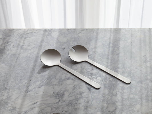 Two stainless steel serving spoons with long handles resting on a gray marble surface in front of white curtains.
