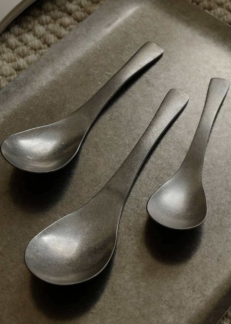 Three matte black metal serving spoons of different sizes arranged diagonally on a dark gray rectangular tray, with a textured beige woven rug visible in the background corner.