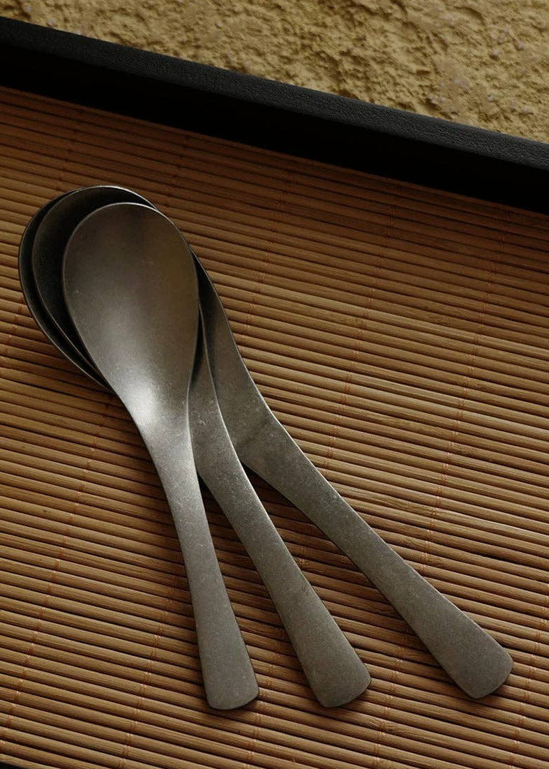 Three metal spoons with a brushed silver finish resting on a brown bamboo placemat.