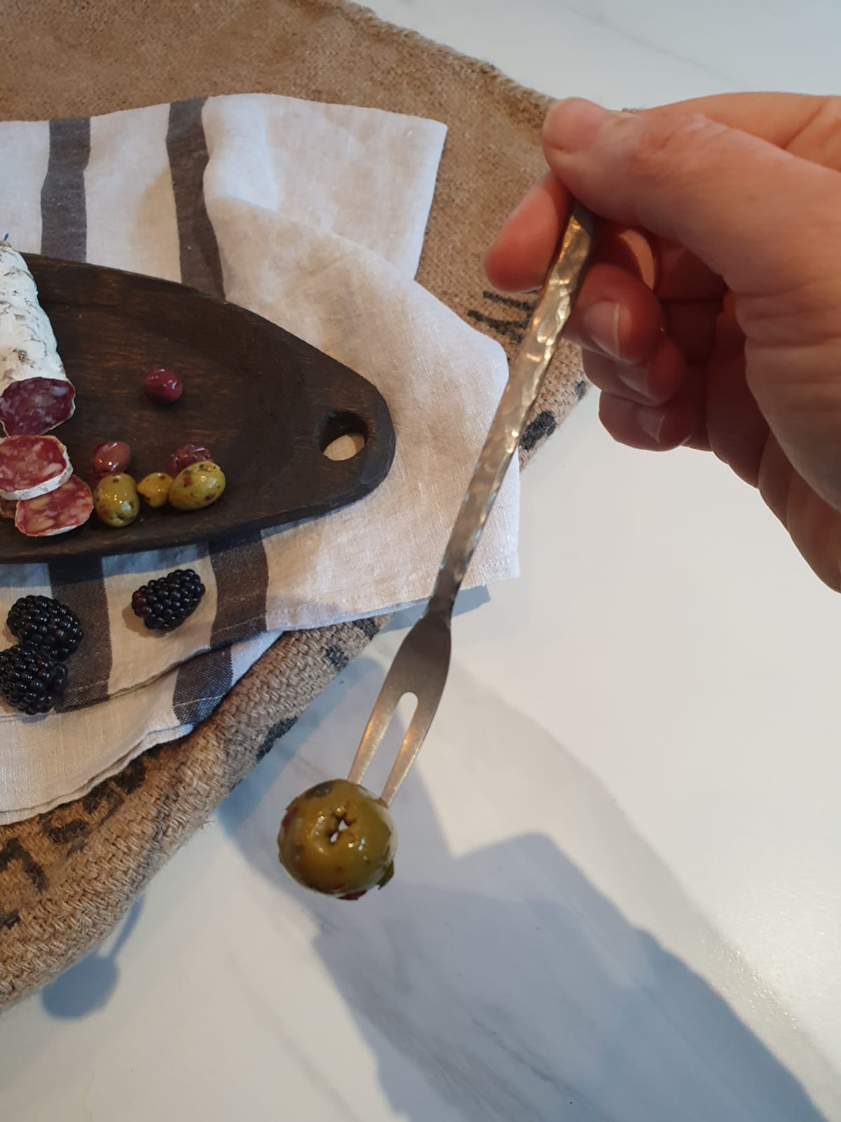 Person holding a fork with an olive on a wooden cutting board with olives and other items.