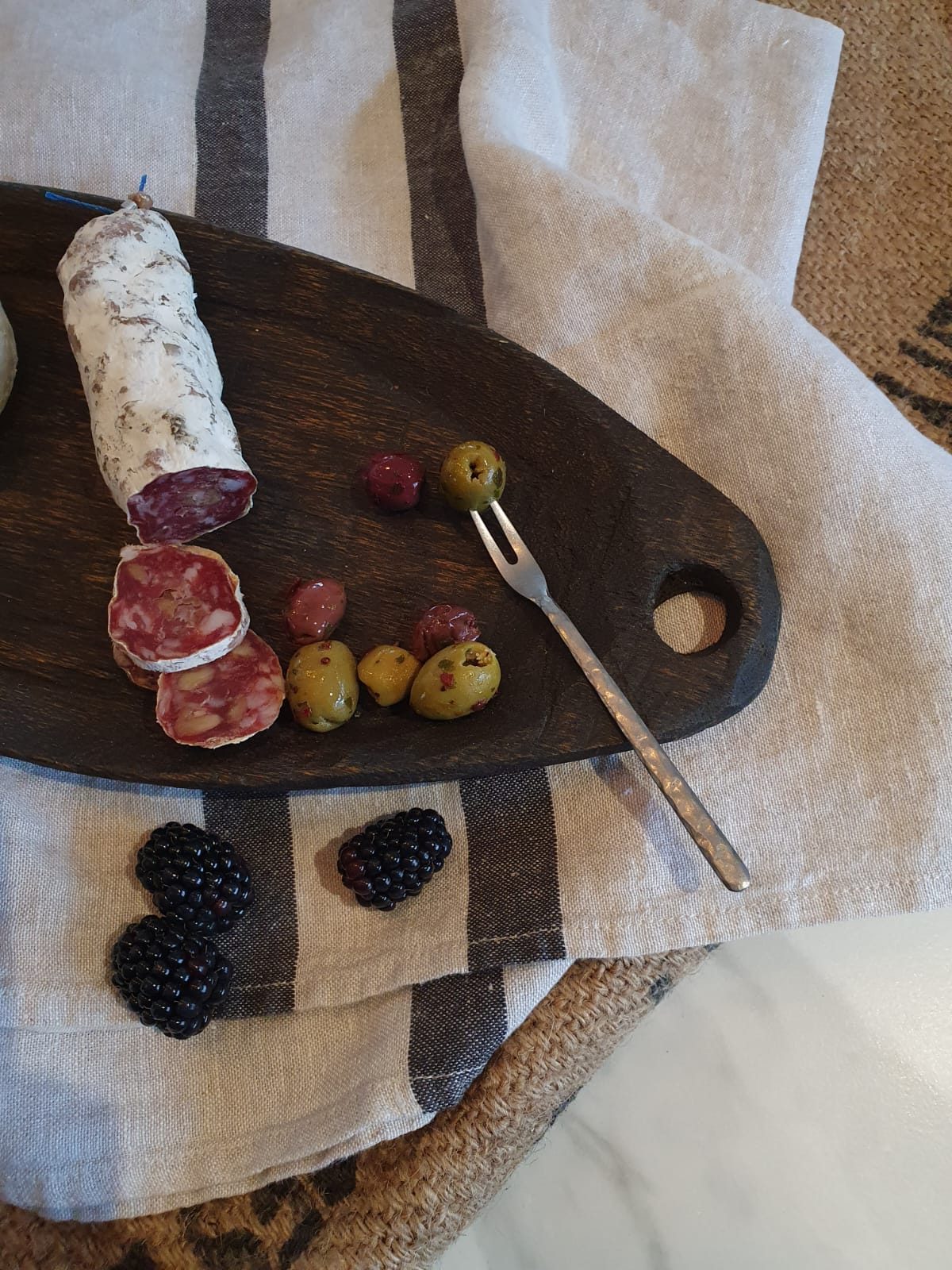 Wooden cutting board with cured meats, olives, and blackberries on a checkered cloth.