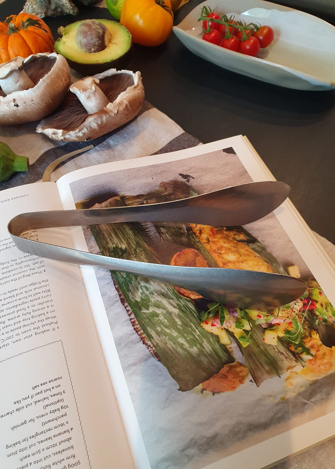 Metal tongs on a magazine with food images, surrounded by vegetables on a table.