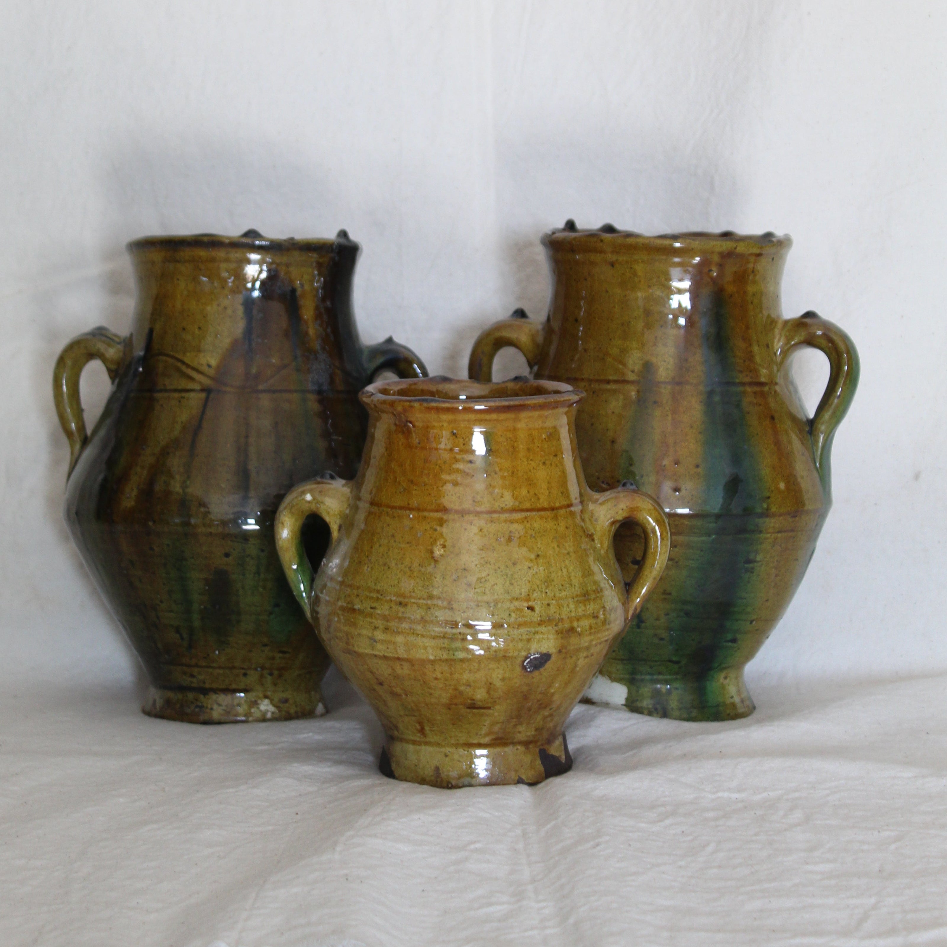 Three handmade ceramic vases with a rustic glaze, displayed on a white fabric surface against a white fabric backdrop.