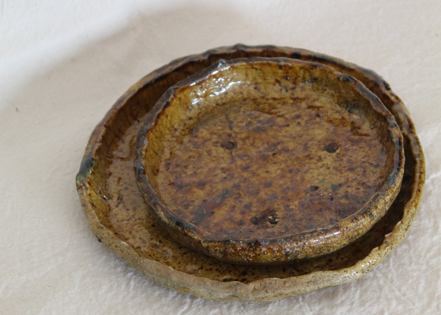 A handmade ceramic dish with a rustic, layered glaze in earthy brown, gold, and green tones, photographed on a textured white fabric background.