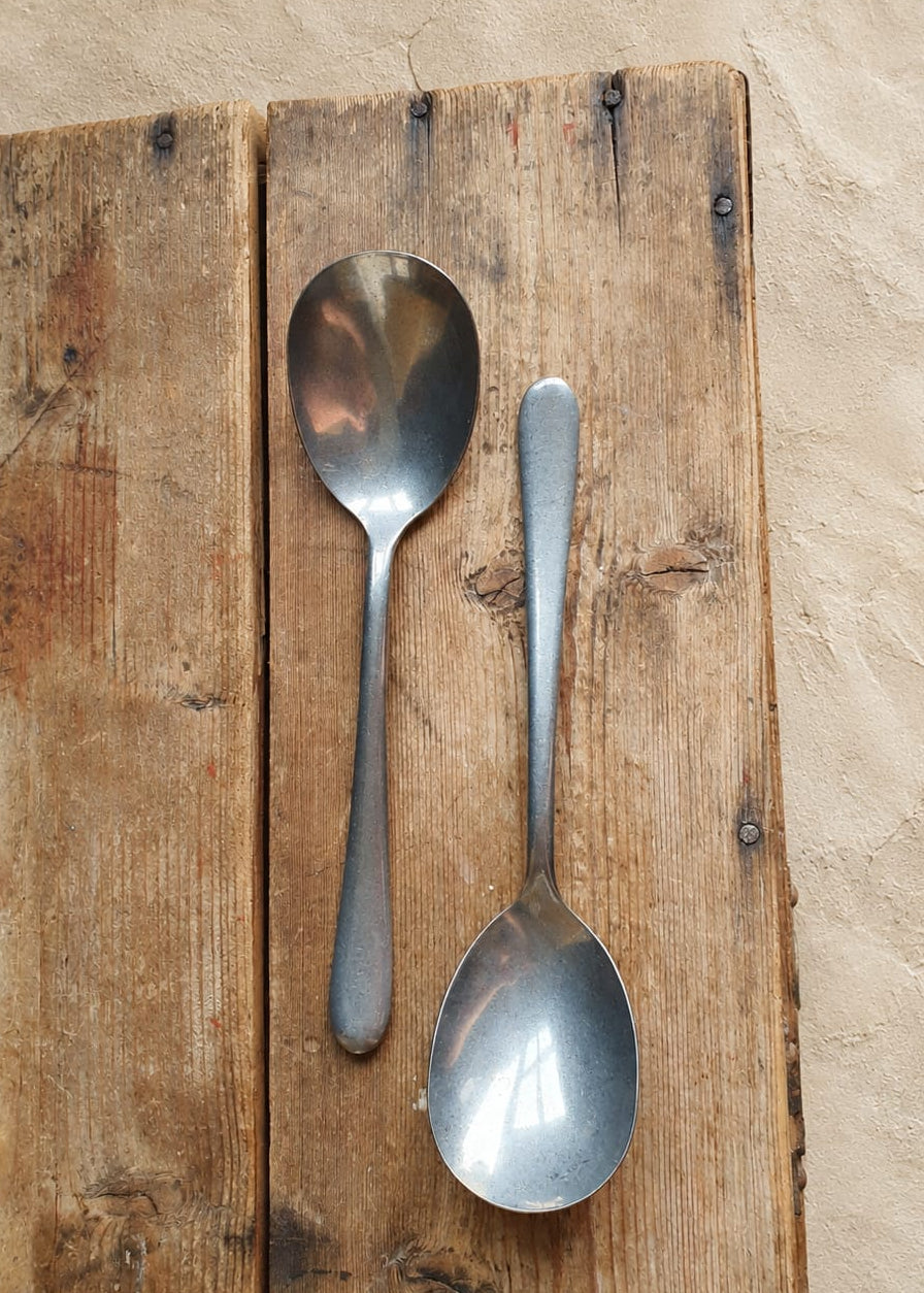 Two vintage silver metal spoons, one larger soup spoon and one smaller serving spoon, displayed vertically on a rustic wooden plank against a textured beige plaster wall.