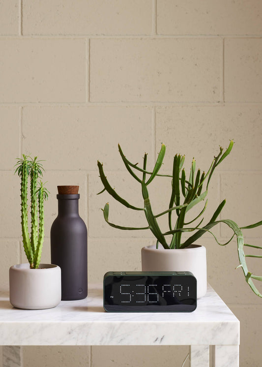 Decorative setup with potted plants and a digital clock on a marble surface against a beige brick wall.